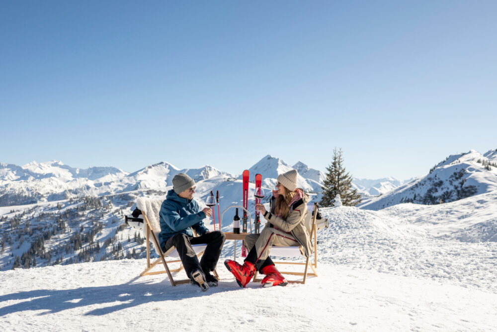 Ski und Weingenusswoche im Großarltal. Zwei Personen in Skibekleidung sitzen auf Liegestühlen und trinken ein Glas Rotwein. Im Hintergrund sieht man die Ski und die Berge.